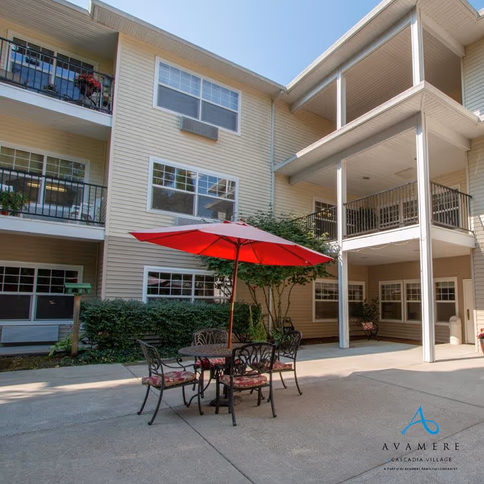 Outdoor courtyard area of a senior living facility with a round metal table and four chairs with cushions under a large red umbrella. The building has beige siding, multiple windows, balconies, and a covered patio area. There are bushes and a small tree near the building.