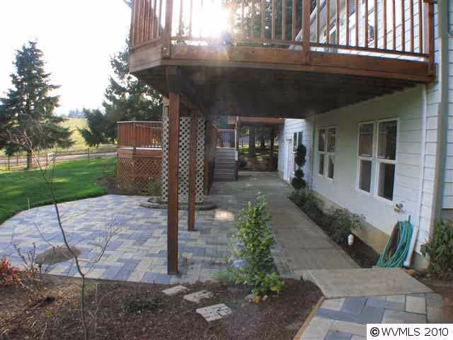 Outdoor patio area with a paved stone floor beneath a wooden deck attached to a white building. There are plants and small trees around the patio, a garden hose coiled near the building, and a grassy area with trees in the background.