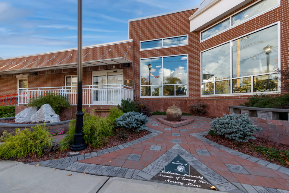 Exterior courtyard and entrance of a brick senior living facility with a paved memorial walkway, landscaping, a lamp post, and large reflective windows.