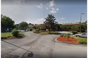 View of the driveway and entrance area of a single-story building with a landscaped roundabout featuring trees and shrubs under a partly cloudy sky.