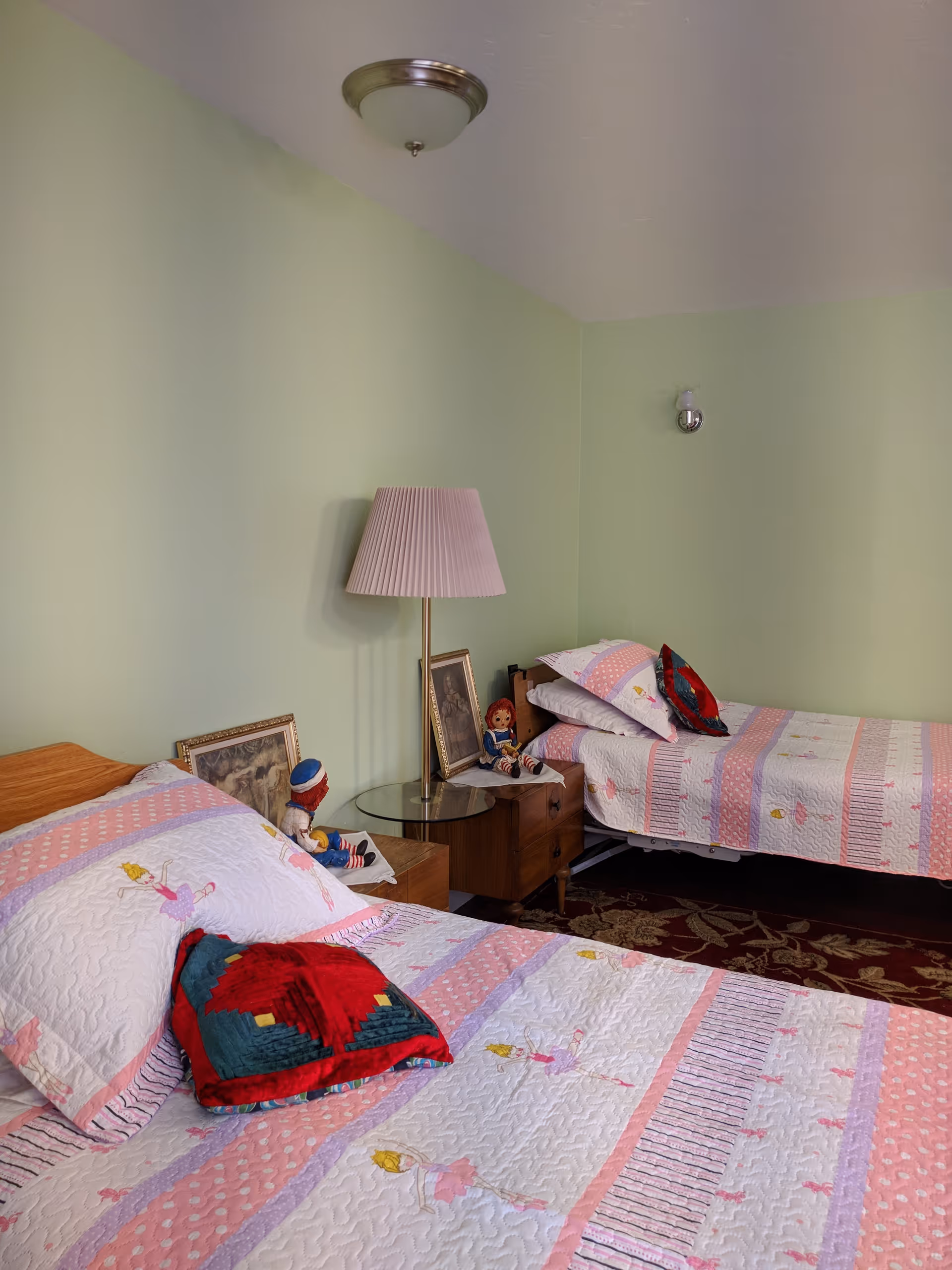 A bedroom with two single beds covered in pink and white patterned quilts featuring ballerina designs. Each bed has a decorative pillow with a red and green pattern. Between the beds is a wooden nightstand with a glass top, a table lamp with a pleated shade, framed pictures, and two Raggedy Ann dolls. The walls are painted light green, and there is a ceiling light fixture and a wall-mounted light fixture above one bed. A patterned rug is partially visible on the floor.