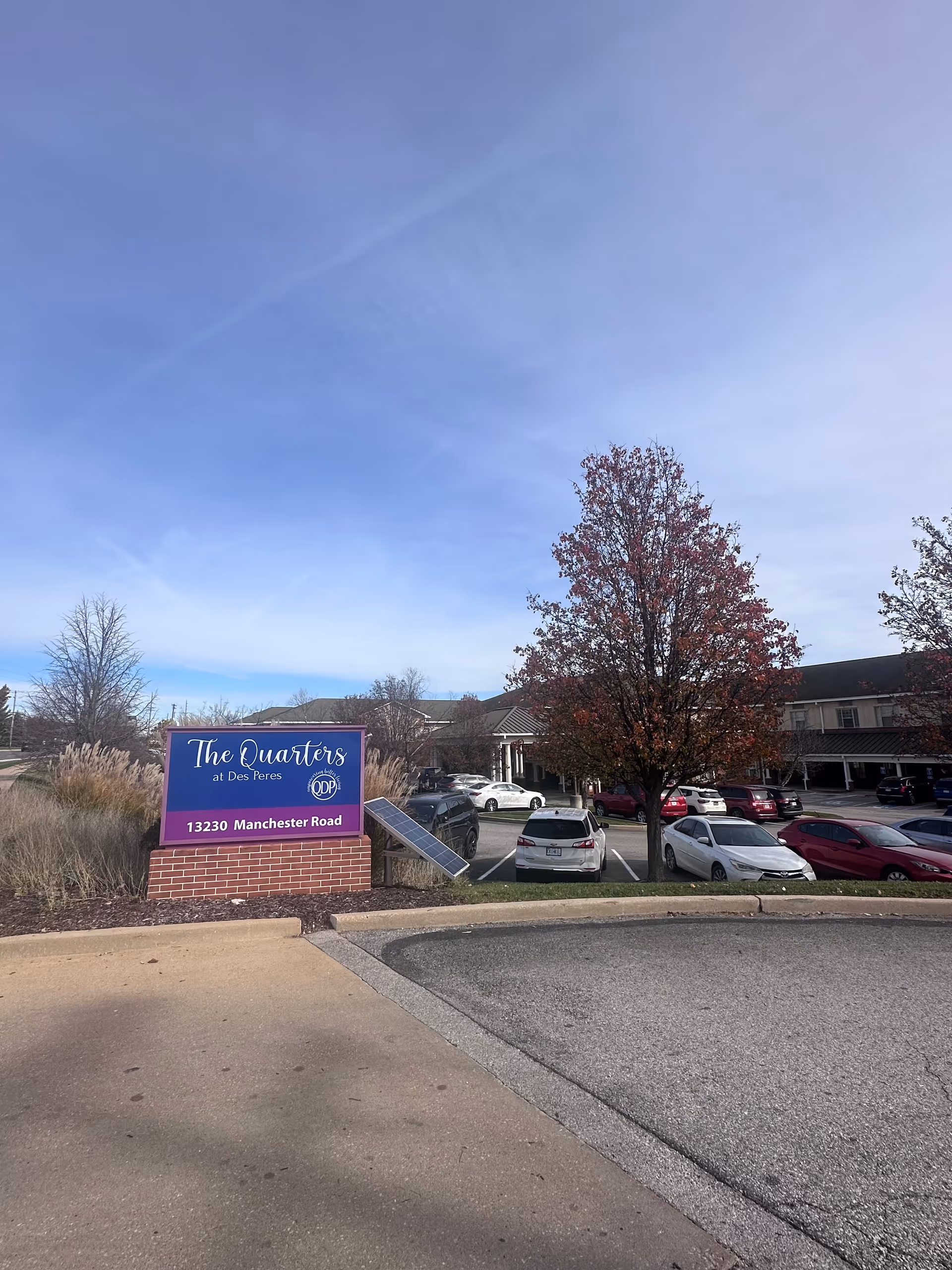 Outdoor view of the parking lot and entrance area of The Quarters at Des Peres facility, with a sign displaying the name and address 13230 Manchester Road. Several cars are parked, and trees with autumn foliage are visible under a clear blue sky.