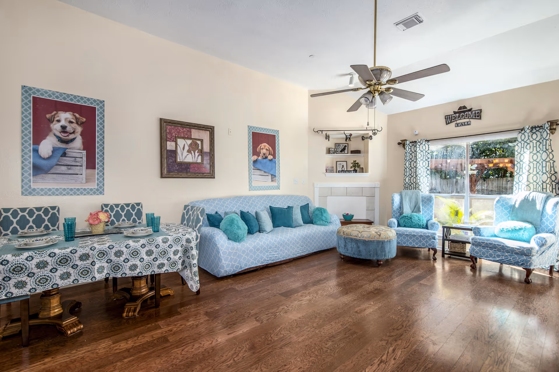 A bright and cozy living room with wooden flooring, a ceiling fan, and large windows with patterned curtains letting in natural light. The room features a light blue patterned sofa with multiple teal and turquoise cushions, two matching armchairs, and a round ottoman. A small dining table with a patterned tablecloth and place settings is visible on the left side. The walls are decorated with framed pictures, including two images of puppies and a floral artwork. A 'WELCOME Y'ALL' sign is mounted above the window.