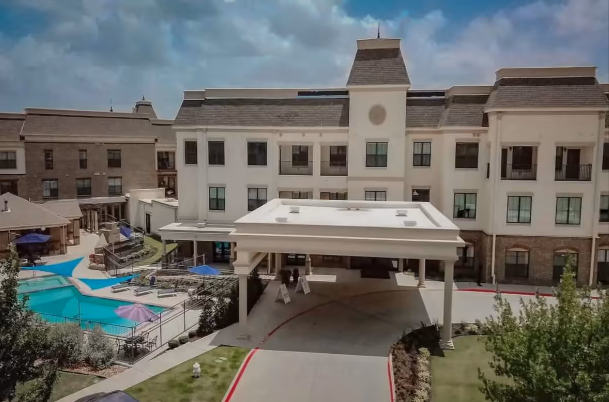 Exterior view of a multi-story senior living facility with a covered entrance driveway, landscaped greenery, and a fenced swimming pool area with umbrellas and lounge chairs under a partly cloudy sky.