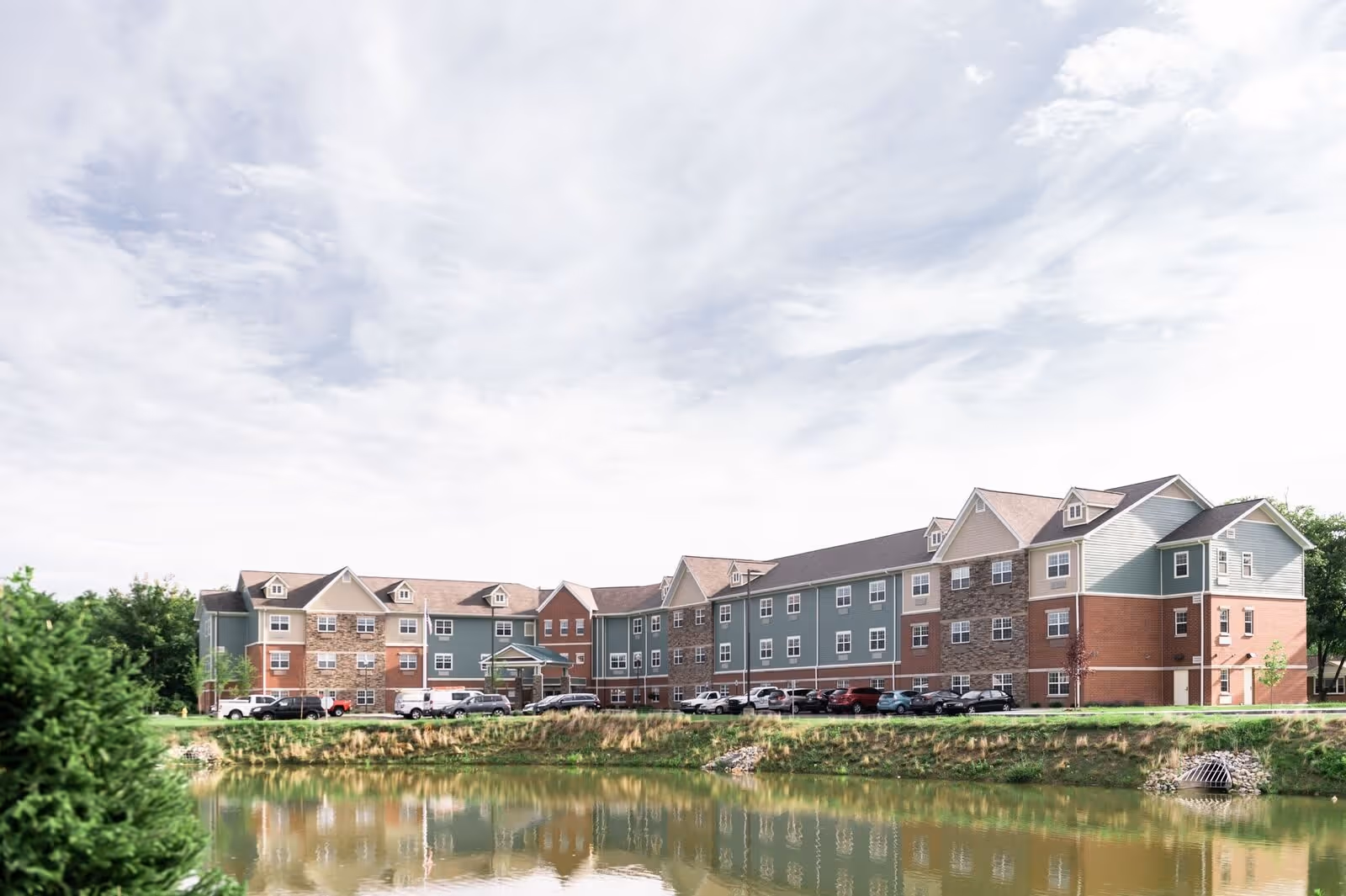 Exterior view of a large three-story senior living facility building with a mix of brick, stone, and siding facade, situated next to a pond with a grassy embankment and trees under a partly cloudy sky.