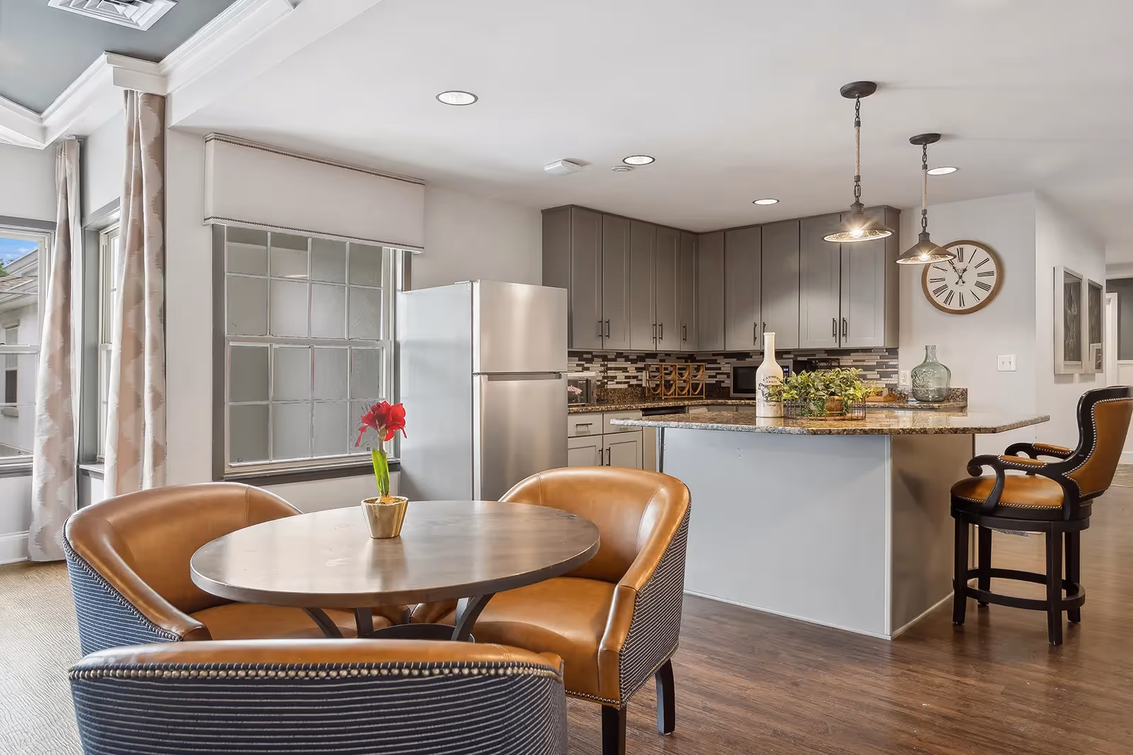 Open-plan kitchen with a granite island, gray cabinets and pendant lights adjacent to a round dining table with leather chairs.