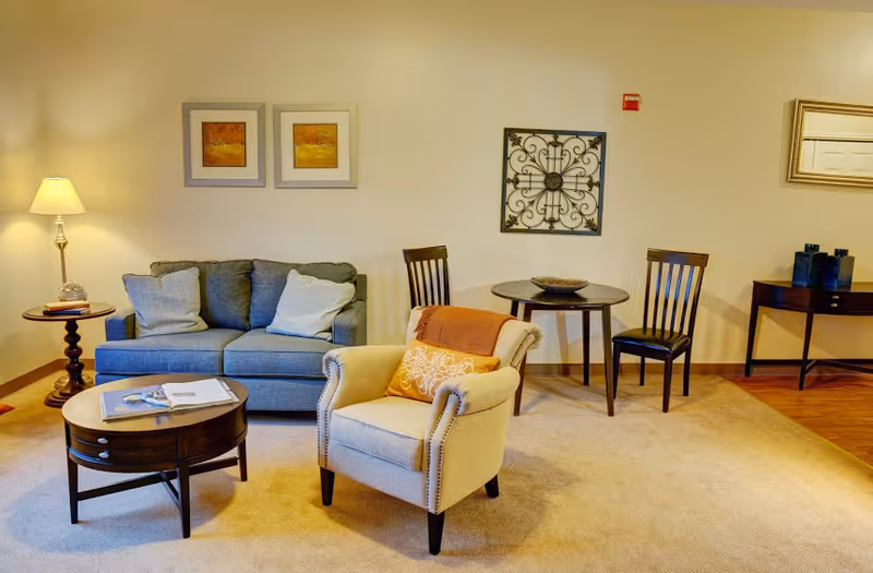 A cozy living room area with a blue sofa adorned with two cushions, a beige armchair with an orange throw and decorative pillow, a round wooden coffee table with magazines, a small round dining table with two wooden chairs, a side table with a lamp, and wall decorations including framed pictures and a decorative metal piece.