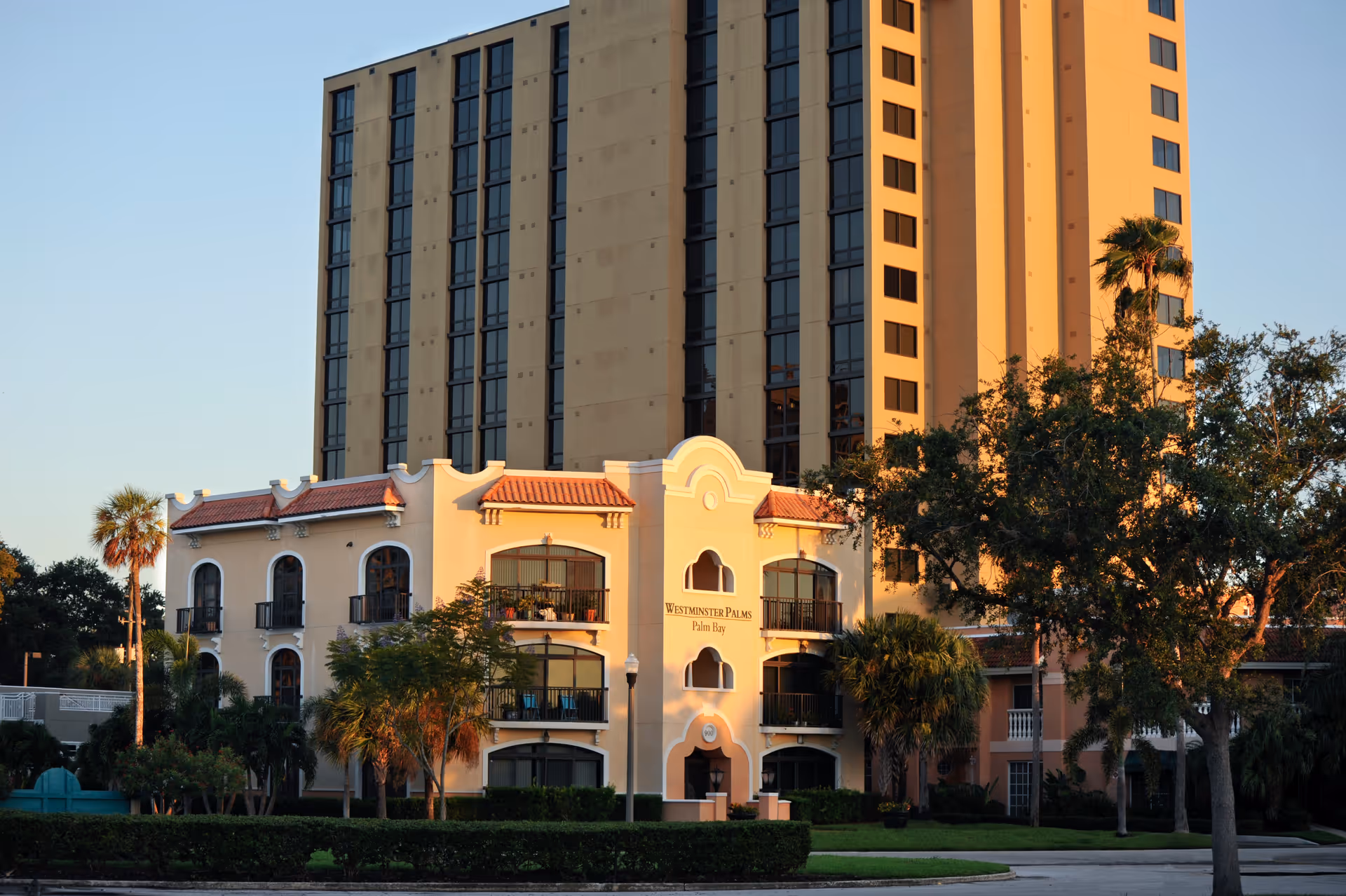 Exterior view of a multi-story senior living facility named Westminster Palms, featuring a beige and light yellow building with balconies, palm trees, and greenery in the foreground during sunset.