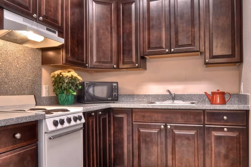 A kitchen with dark wooden cabinets, a white electric stove with four burners, a microwave on the countertop, a small sink, a red teapot, and a potted plant with yellow flowers.