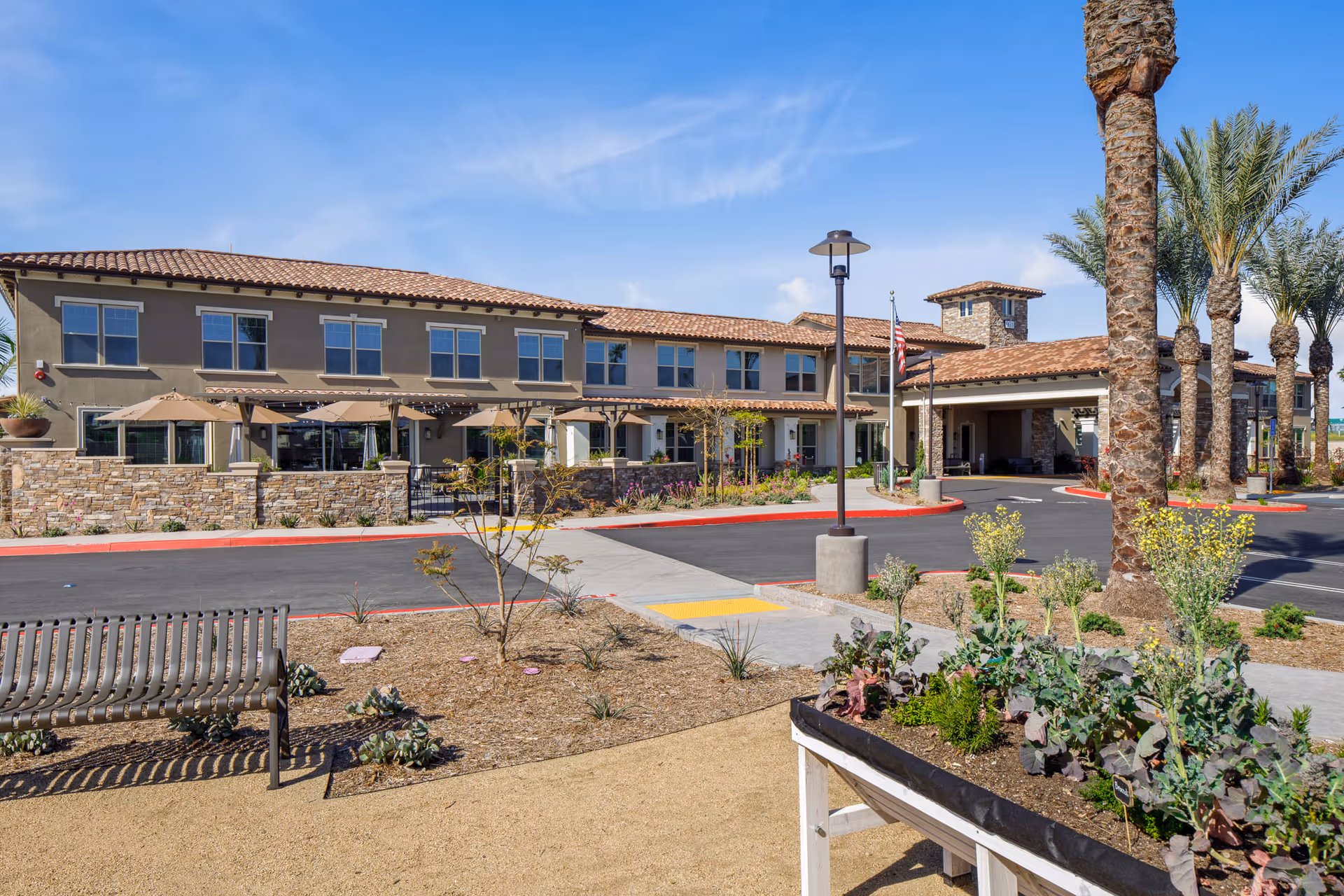 Exterior view of Oakmont of Riverpark senior living facility showing a two-story building with a tiled roof, outdoor seating area with umbrellas, palm trees, landscaped garden beds, a bench, and a driveway with a covered entrance under a clear blue sky.