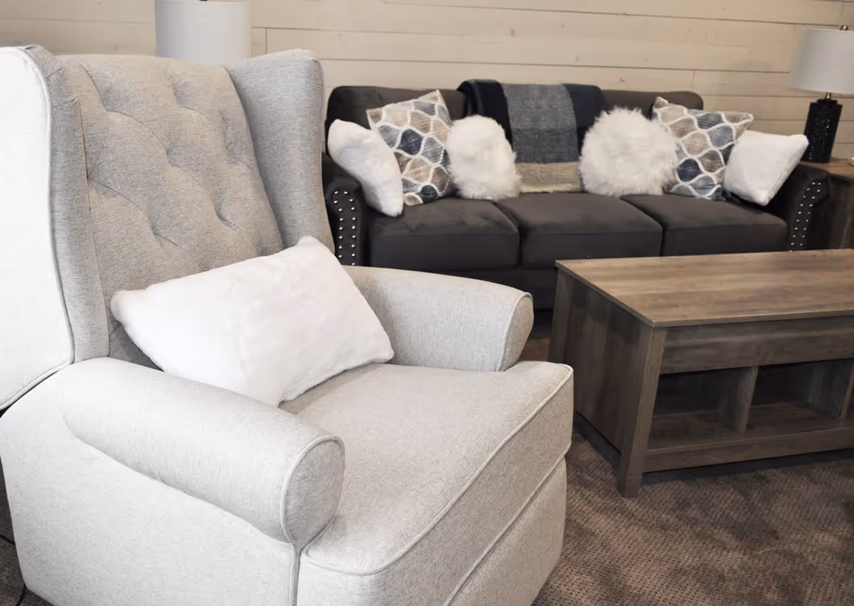 A cozy living room area featuring a light gray upholstered armchair with a white pillow, a dark gray sofa adorned with patterned and fluffy white pillows, a wooden coffee table, and a side table with a black lamp. The background has light-colored wooden paneling on the wall.