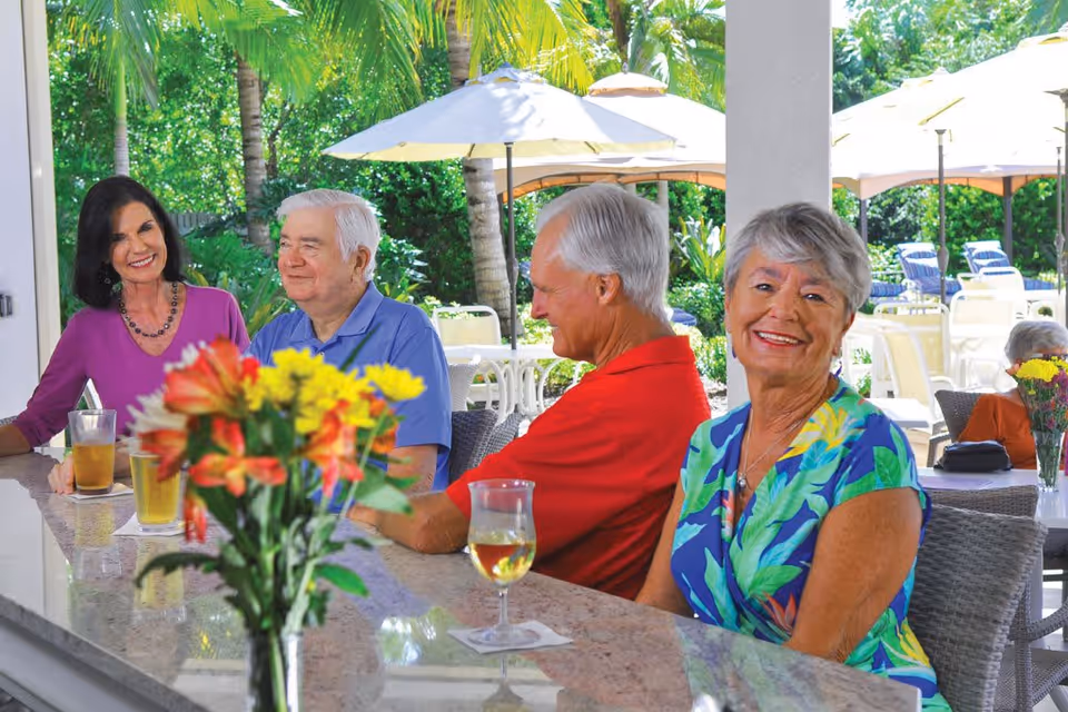 Four elderly people sitting at an outdoor bar counter with drinks, smiling and enjoying a sunny day. Behind them are palm trees, umbrellas, and patio furniture in a garden-like setting.