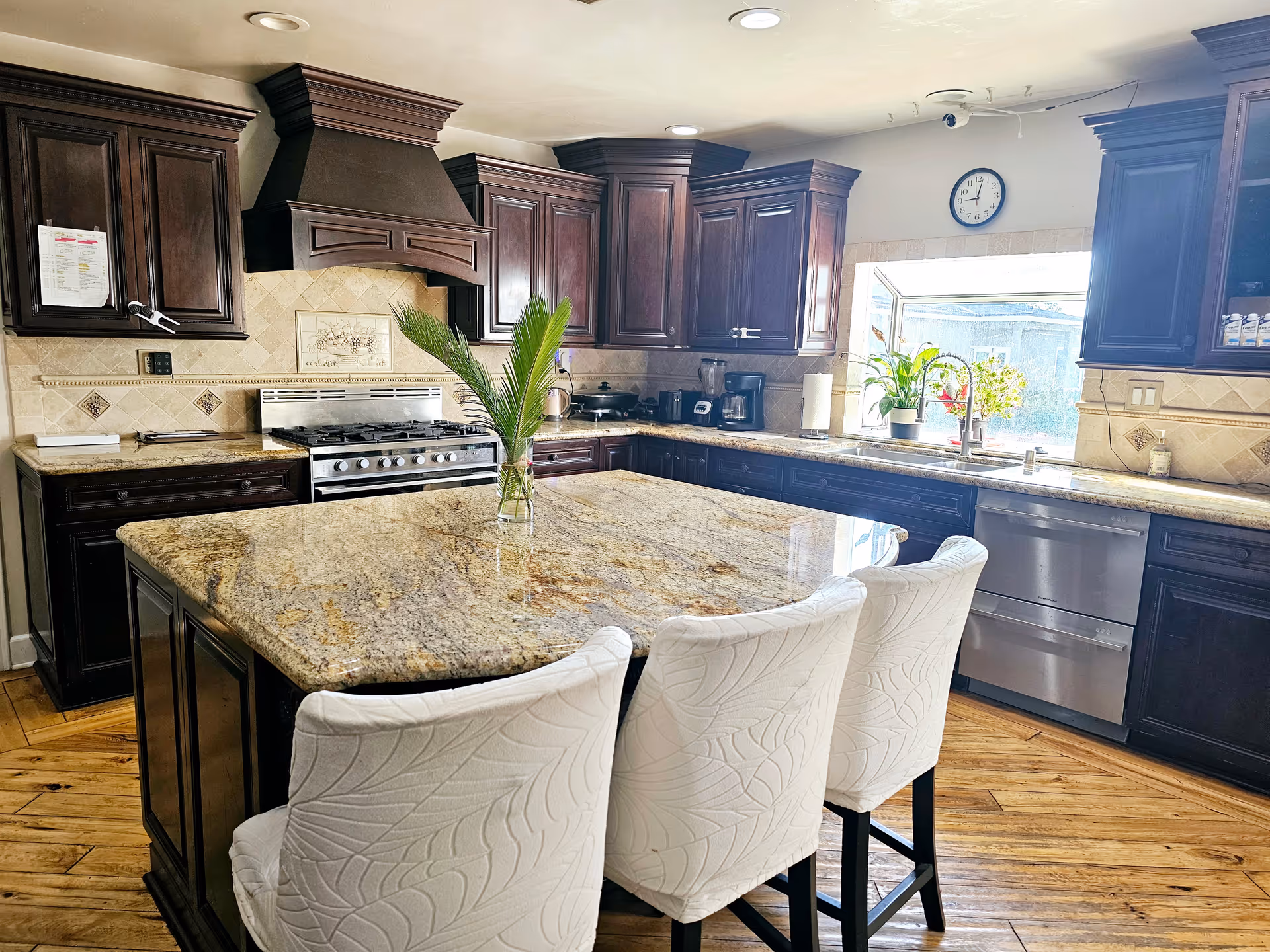 A spacious kitchen with dark wooden cabinets, a large granite island with three white cushioned chairs, stainless steel appliances including a stove and dishwasher, a window above the sink with plants on the windowsill, and a clock on the wall.