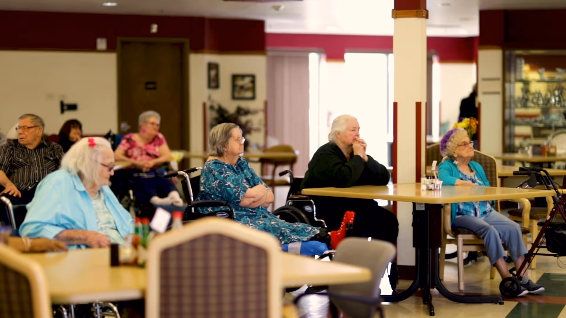 Several elderly residents seated around tables in a communal dining/activity room at a senior living facility.
