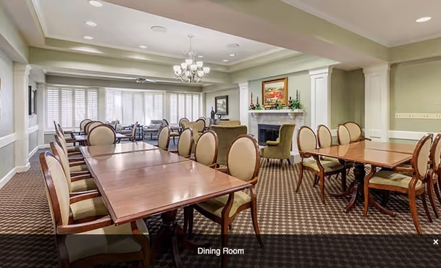 A spacious dining room with multiple wooden tables and cushioned chairs arranged neatly. The room features a chandelier, a fireplace with a painting above it, and large windows with white shutters allowing natural light to fill the space. The carpet has a patterned design, and the walls are painted in a light green color.
