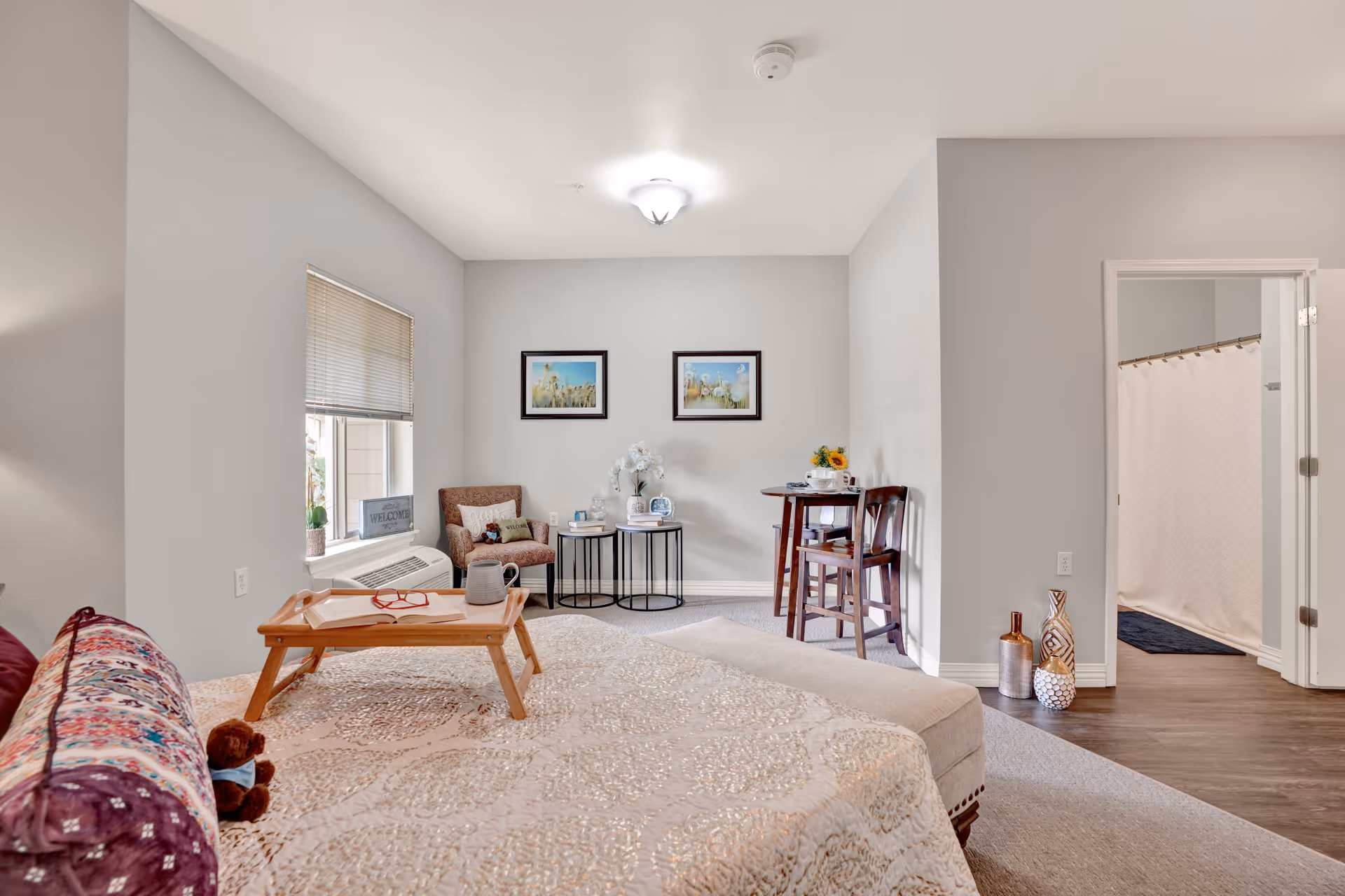 A cozy bedroom in a senior living facility with a bed covered in a patterned quilt and a small wooden tray on top. There is a window with blinds, a cushioned chair, two framed pictures on the wall, a small round table with flowers and books, and a small dining table with two chairs. An open door reveals a bathroom with a shower curtain.