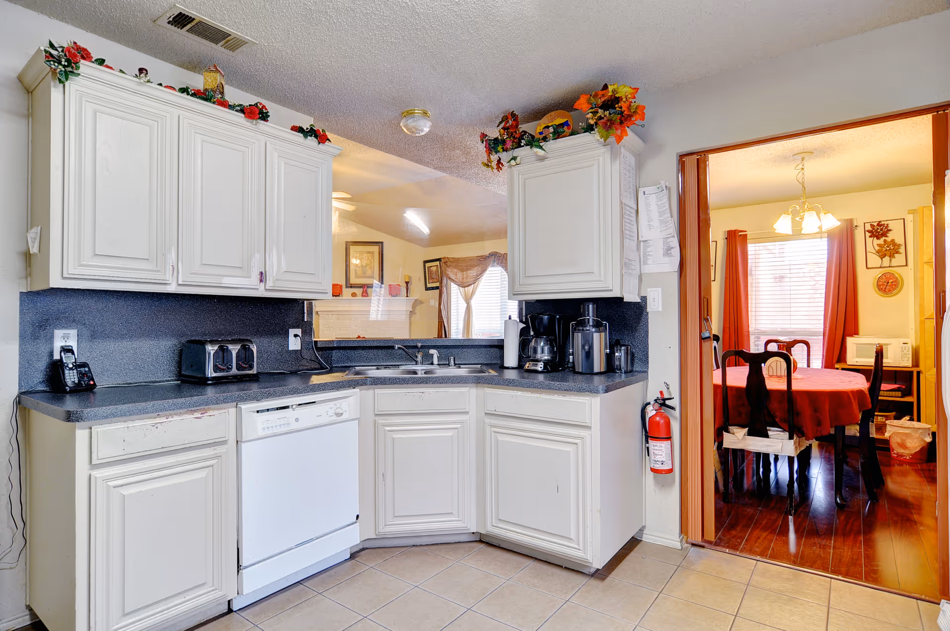 Bright kitchen with white cabinets, black countertops, a sink and appliances, opening into a dining area with a red-tableclothed table.
