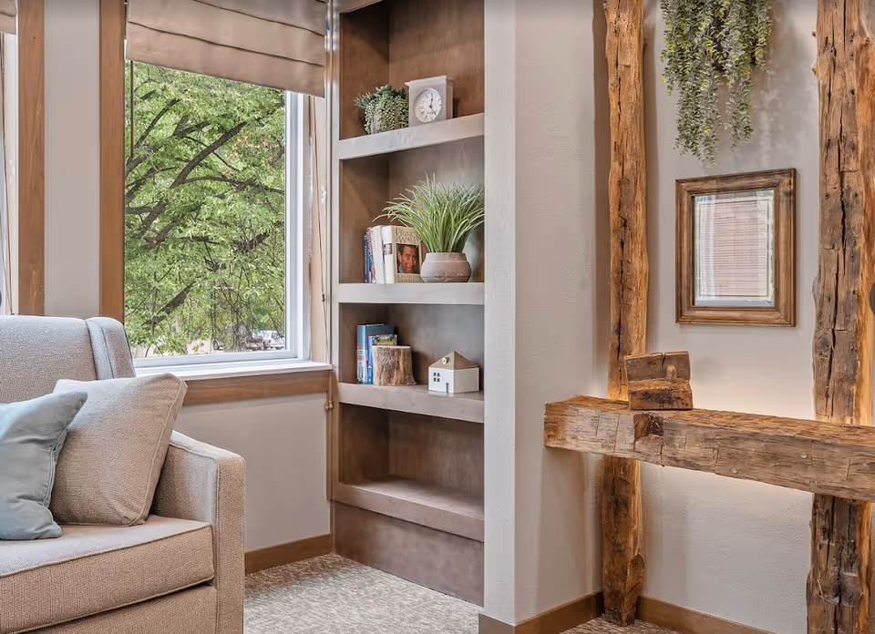 Cozy sitting area with an upholstered armchair by a window, built-in shelves with books and plants, and rustic wooden beams and decor on the wall.