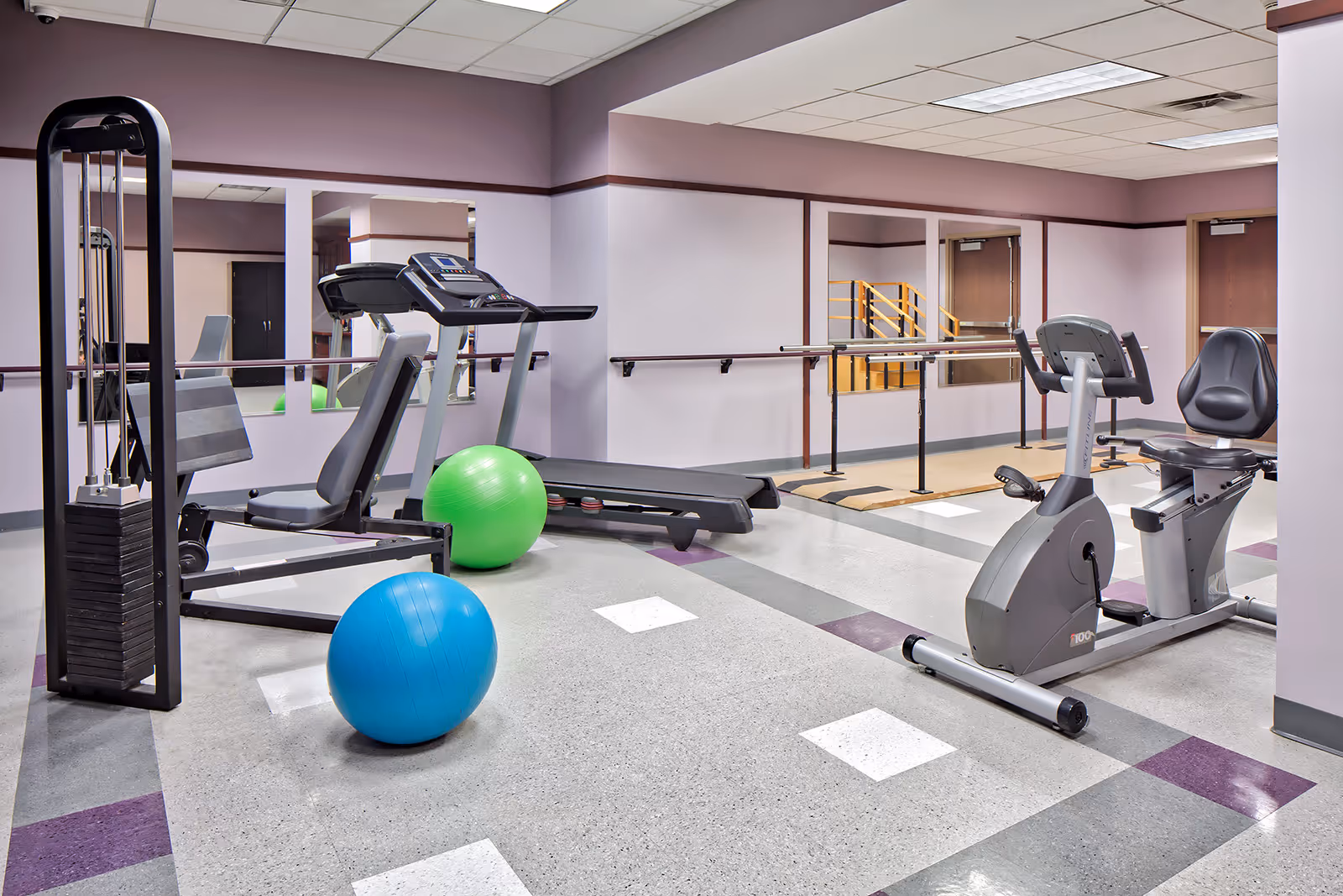 A fitness room in Clark Manor featuring exercise equipment including a recumbent stationary bike, a treadmill, a weight machine, and two large exercise balls on a tiled floor with purple and gray accents. The room has light purple walls with mirrors and handrails along the walls.