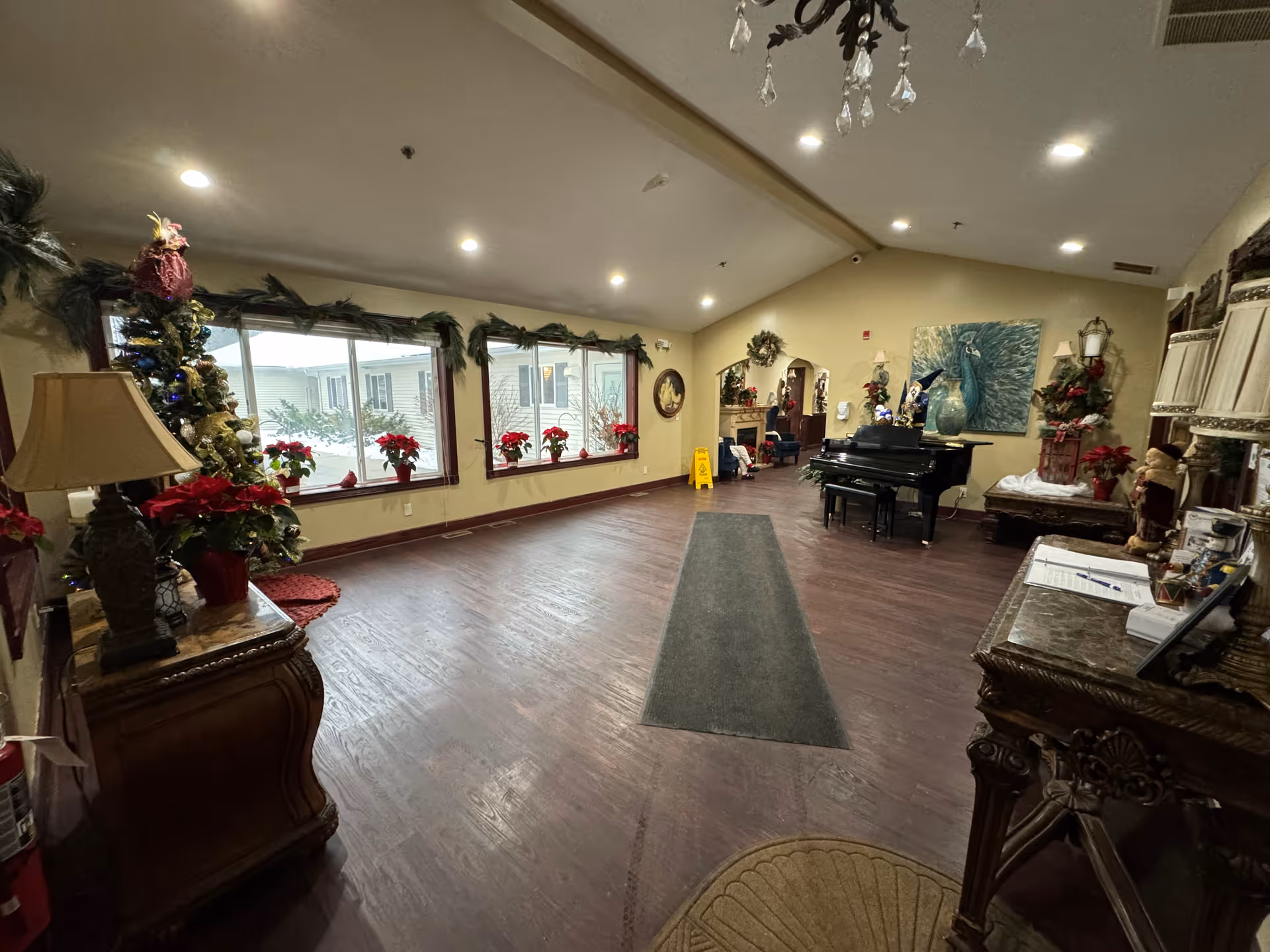 Interior view of a spacious assisted living facility common area with wooden floors, large windows decorated with garlands and poinsettia plants, a black grand piano, and various decorative furniture including lamps and a side table. The room is warmly lit with ceiling lights and has holiday decorations.