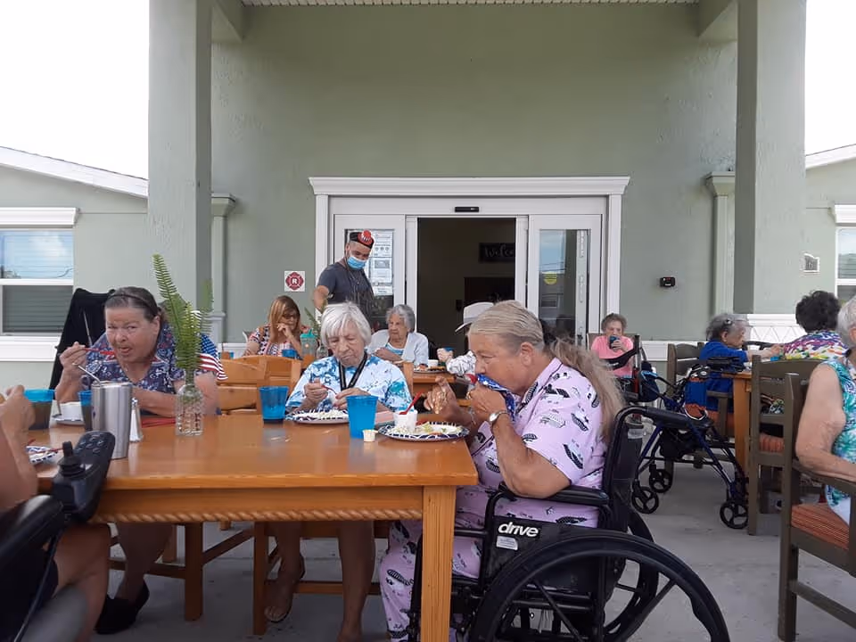 A group of elderly people sitting around wooden tables outdoors at a senior living facility, eating and socializing. Some are in wheelchairs, and a staff member wearing a mask is standing near the entrance of the building in the background.
