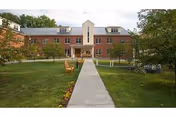 A three-story brick building with a central entrance, surrounded by a well-maintained lawn and trees. A concrete walkway leads from the foreground to the entrance, with benches and flower beds along the path.