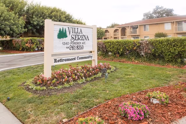 Entrance sign for Villa Serena Retirement Community on a landscaped lawn with flowers and the apartment building in the background.