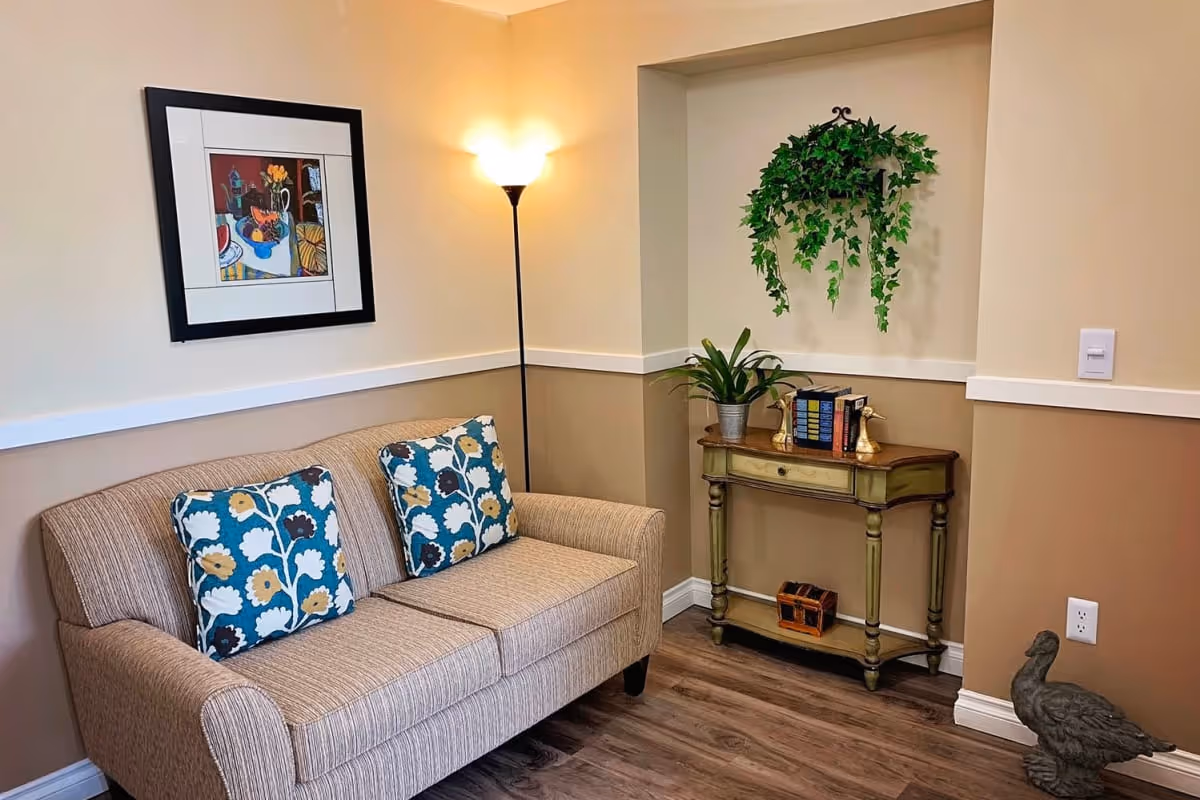 A cozy living room corner with a beige striped sofa adorned with two floral patterned cushions. A tall floor lamp with a glowing light stands behind the sofa. On the adjacent wall, there is a framed colorful artwork. A small wooden console table holds a potted plant, a set of books, and two decorative bird figurines. Above the table, a hanging green leafy plant decorates a recessed wall niche. The floor is wooden, and a small duck statue is placed near the wall outlet.