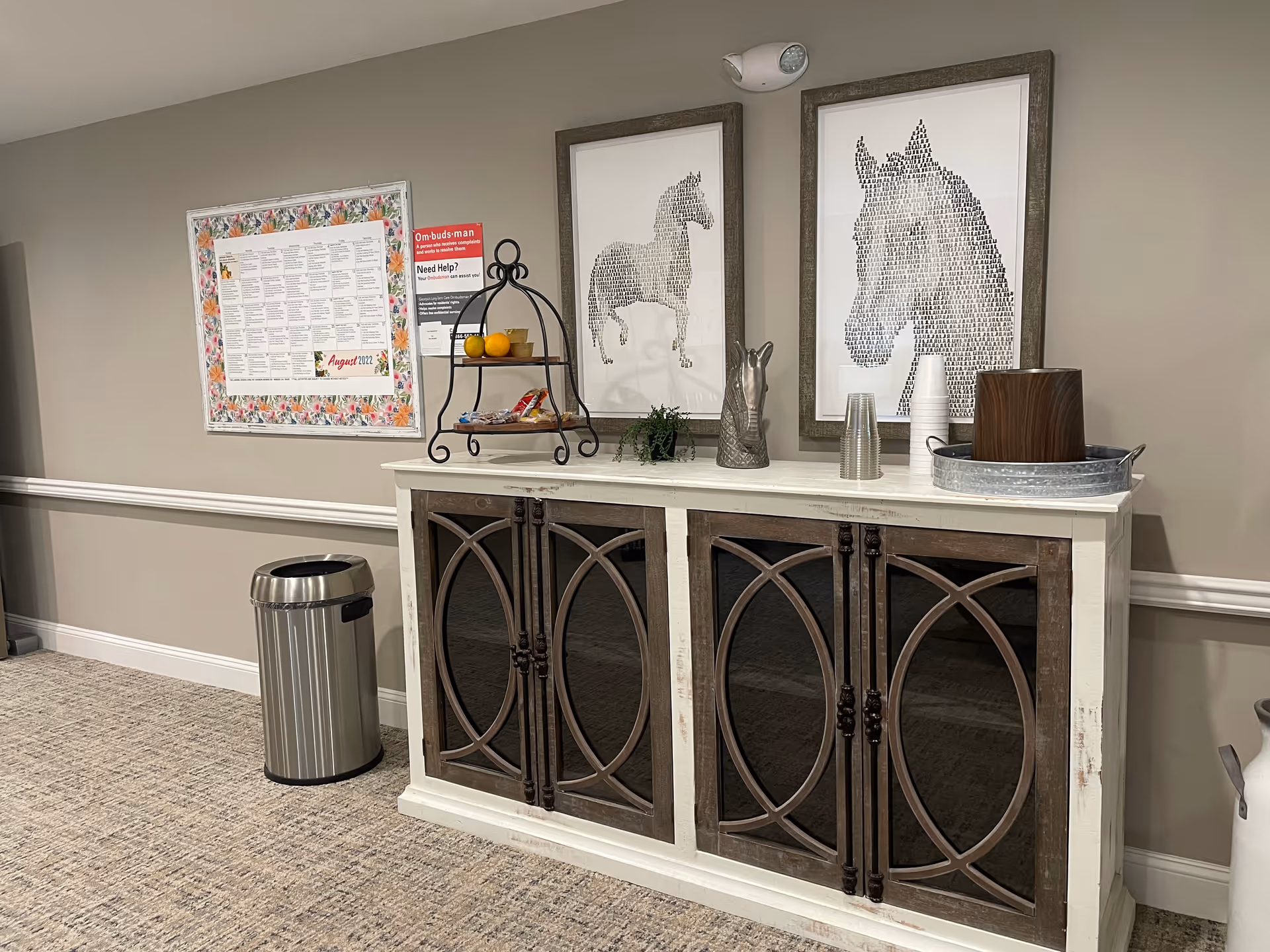 Interior hallway area with a decorative cabinet featuring dark wood and glass doors. On top of the cabinet are two framed horse silhouette artworks, a tiered stand with snacks and fruit, a small plant, a horse head sculpture, plastic cups, and a container with a lid. A stainless steel trash can is positioned to the left of the cabinet, and a bulletin board with notices is mounted on the wall above it.