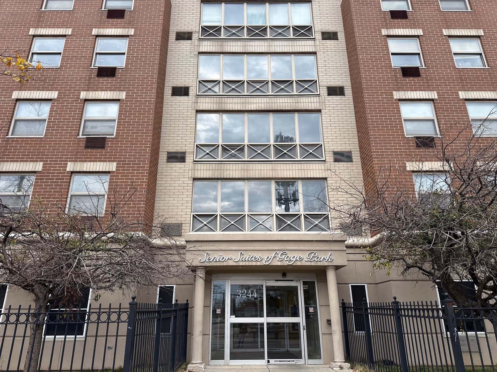 Front exterior view of a multi-story senior living facility named Senior Suites of Gage Park, with a beige and red brick facade, multiple windows, a gated entrance, and the address number 3244 above the glass doors.