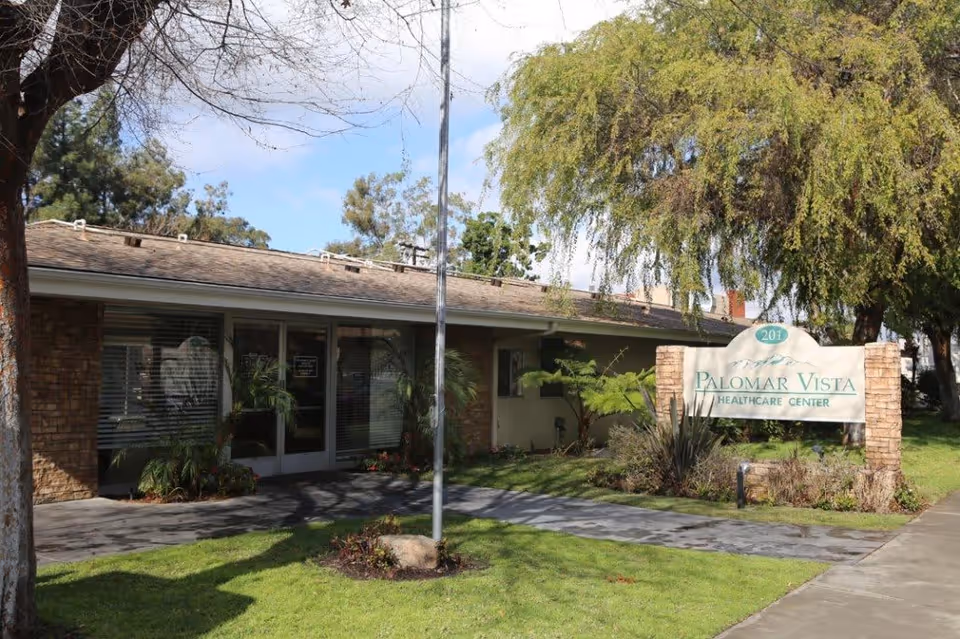 Exterior view of Palomar Vista Healthcare Center building with a sign displaying the facility name and address number 201. The building has a low roof, large windows, and is surrounded by trees and green grass.