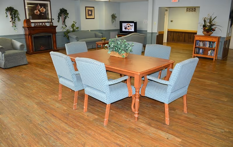 A spacious common area with a wooden table surrounded by six blue upholstered chairs. In the background, there are two sofas, a television, a wooden fireplace mantel with decorative items, framed pictures on the walls, and several potted plants. The floor is wooden and the walls are painted light blue with a chair rail molding.