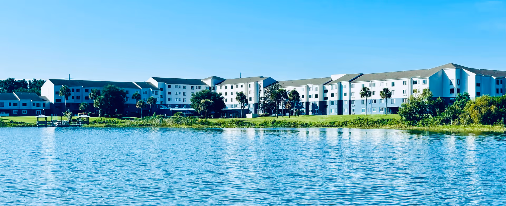 Wide exterior view of a large multi-story senior living facility named Spring Haven, situated beside a calm body of water with green landscaping and palm trees under a clear blue sky.