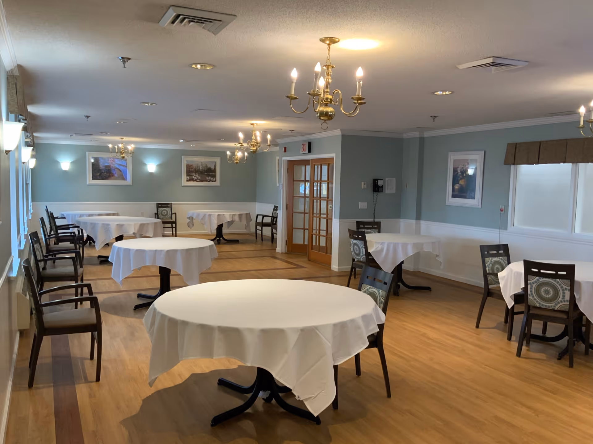 A dining room in Mount Carmel Care Center with round tables covered in white tablecloths and wooden chairs with patterned cushions. The room has light blue walls, wooden flooring, framed pictures on the walls, and brass chandeliers hanging from the ceiling. There are windows along one side and double wooden doors at the far end.