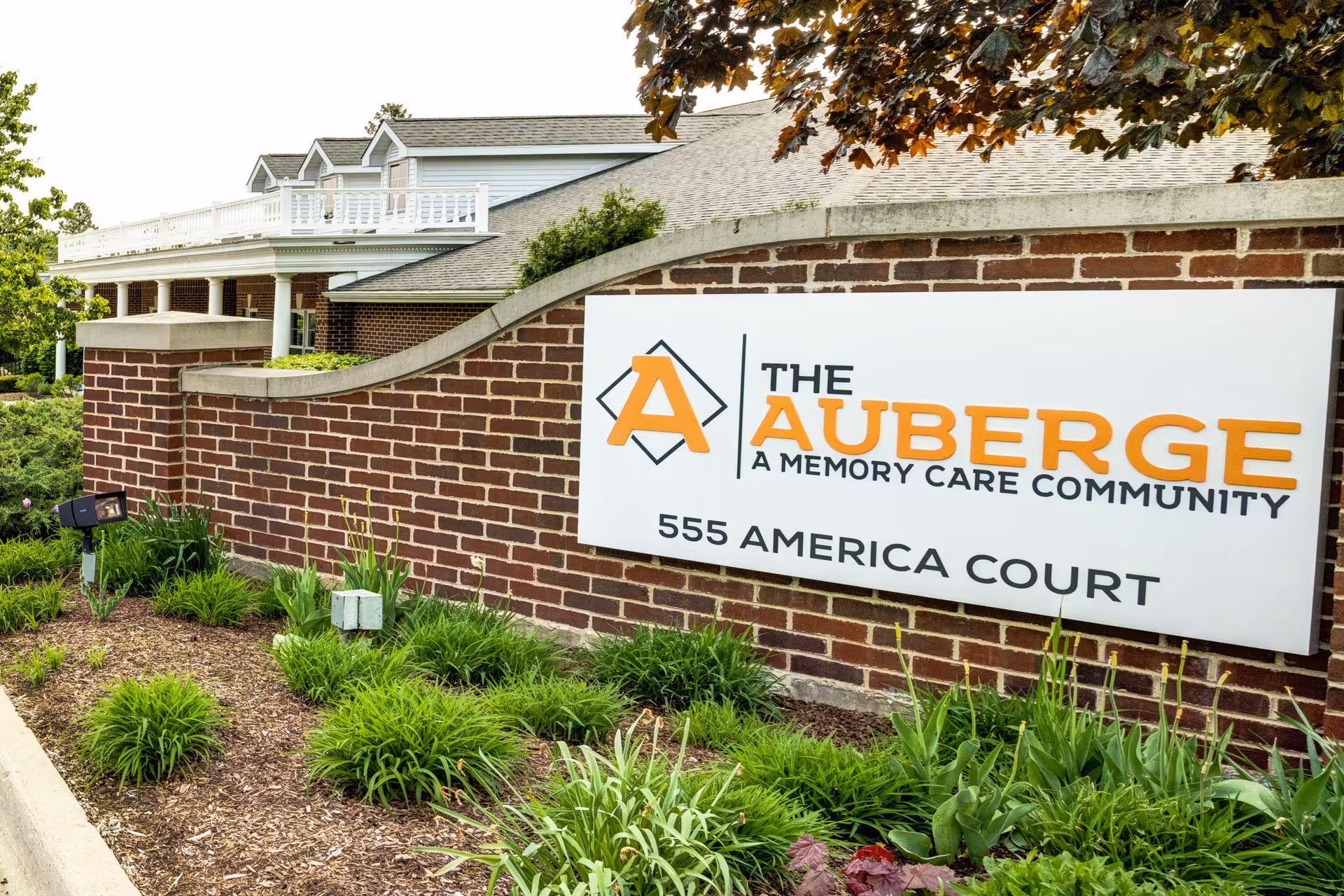 Exterior view of a brick wall with a large sign for The Auberge, a memory care community, located at 555 America Court. The building and greenery are visible in the background.
