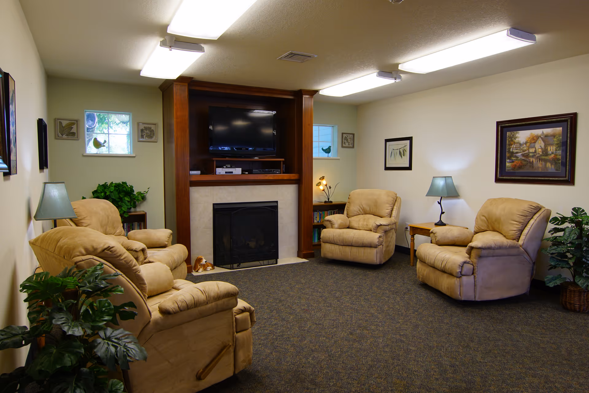Cozy common room with four beige recliners arranged around a fireplace and wall-mounted TV, accented by lamps, plants, and framed artwork.