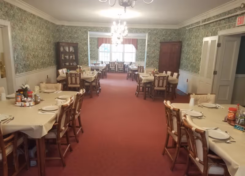 Long dining room with multiple tables set for meals, chairs, chandeliers, patterned wallpaper and a large window at the far end.