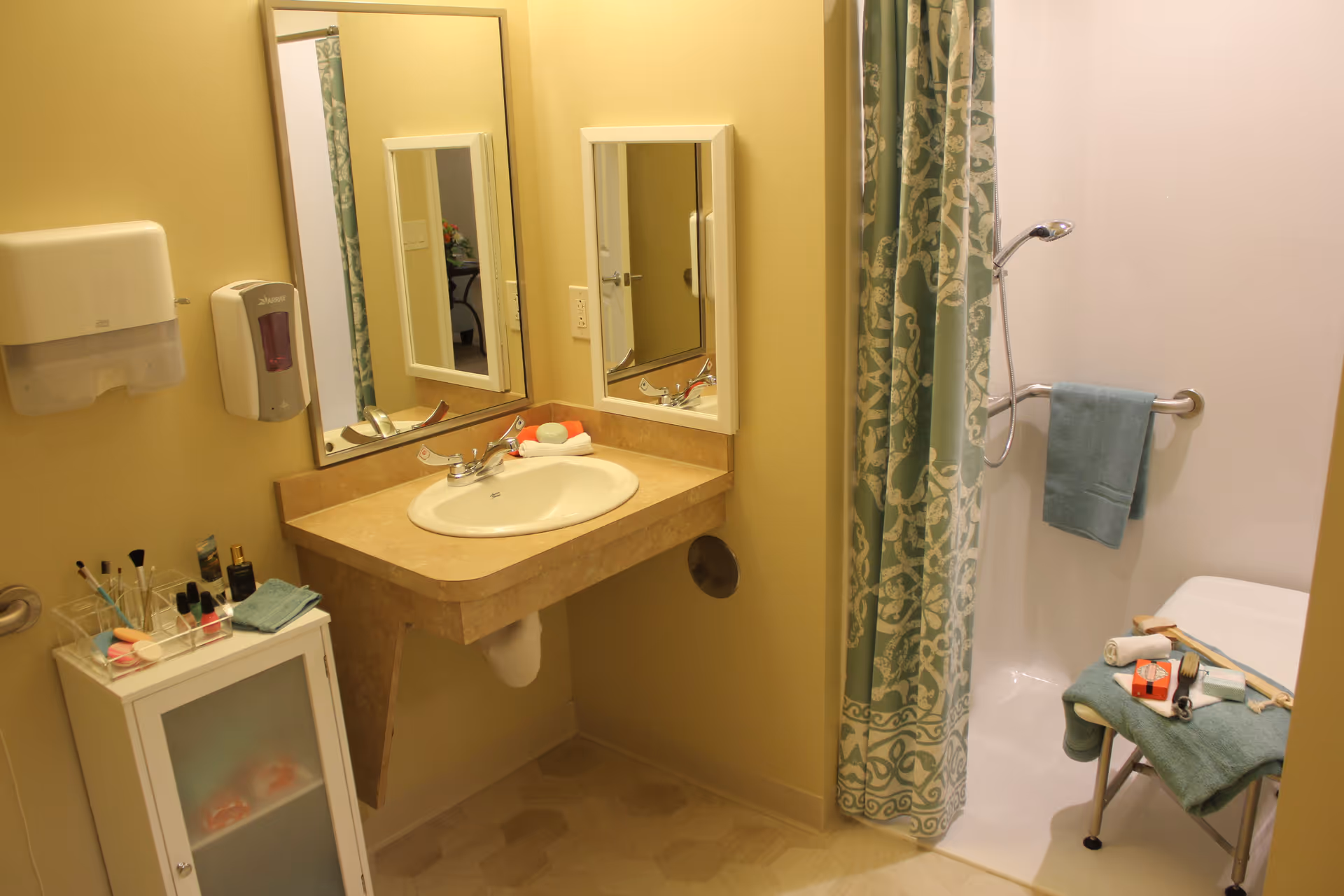 Bathroom with a sink and vanity, wall mirror, and a shower with a patterned curtain, grab bar and shower chair.