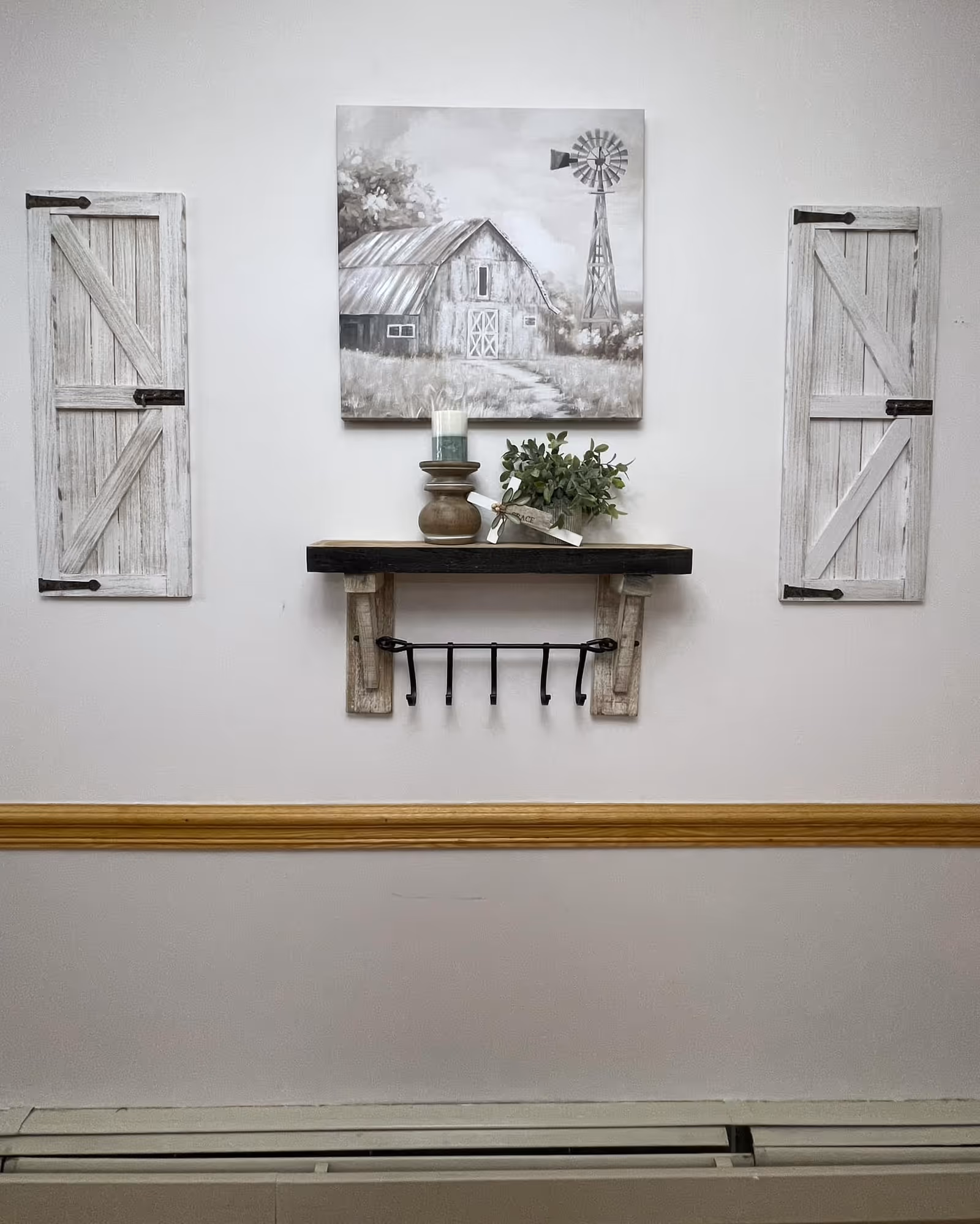 A wall decorated with a rustic wooden shelf holding a candle and a small plant. Above the shelf is a painting of a barn and windmill in a grayscale color scheme. On either side of the painting are two decorative wooden shutters with black metal hinges. Below the shelf is a black metal rack with hooks. The wall has a wooden chair rail molding and a baseboard heater at the bottom.