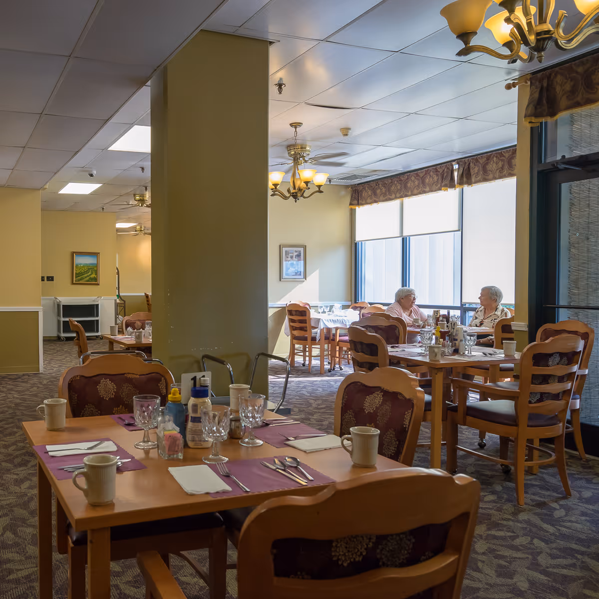 Dining room in a senior living facility with wooden tables and chairs. Tables are set with placemats, utensils, glasses, and condiments. Two elderly women are seated at a table near large windows with blinds, engaged in conversation. The room has carpeted floors, beige walls, ceiling fans, and chandeliers.