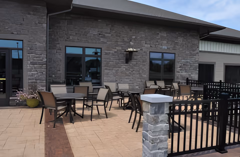 Outdoor patio area with multiple tables and chairs arranged on a paved surface next to a building with stone walls and several windows under a clear blue sky.