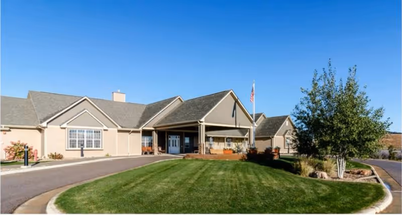Exterior view of Aspen Grove Assisted Living facility showing a single-story building with a covered entrance, a well-maintained lawn, a tree, and an American flag on a flagpole under a clear blue sky.