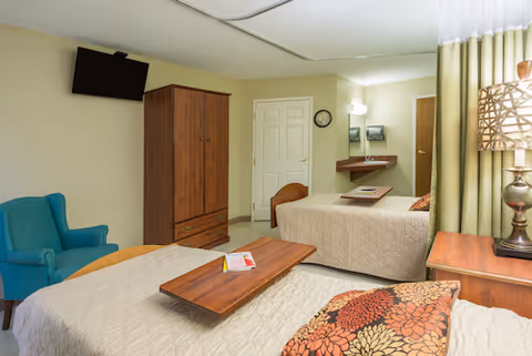 A shared senior bedroom with two beds, wooden bedside tables, a wall-mounted TV, an armoire, and an upholstered armchair.
