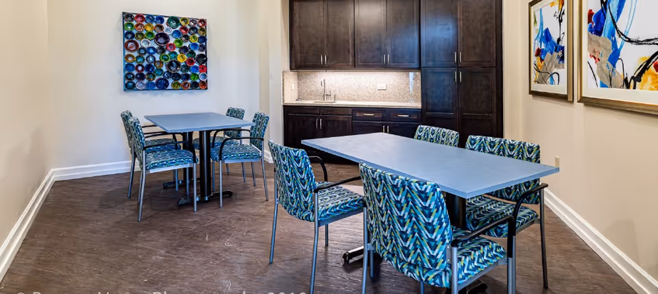 Interior room with two rectangular tables surrounded by patterned chairs. The room has dark wood cabinets with a countertop and sink along one wall. Colorful abstract artwork is displayed on the walls.
