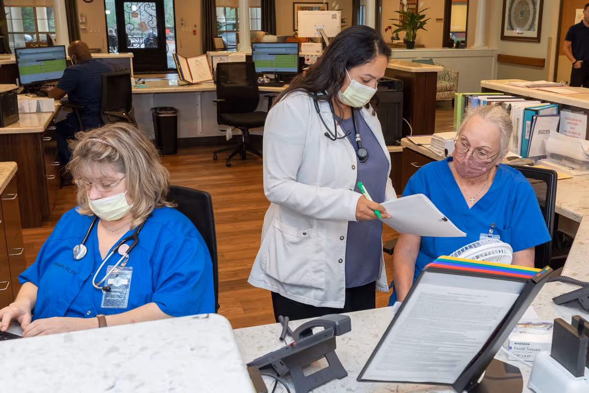 Three healthcare workers wearing masks are at a nursing station inside a medical facility. Two women in blue scrubs are seated, one looking at a phone and the other looking at documents, while a woman in a white coat stands between them, holding a clipboard and pen. Computer monitors and office supplies are visible in the background.