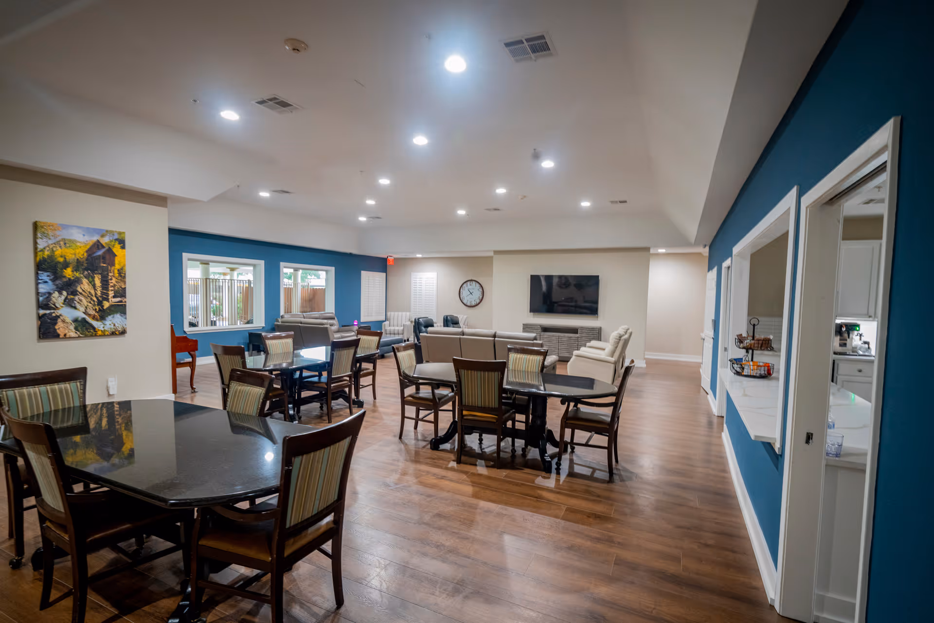 A spacious senior living common area with multiple tables and chairs arranged for dining or socializing. The room features wood flooring, a blue accent wall with two pass-through windows to a kitchen area, a wall-mounted TV, a clock, and comfortable seating including sofas and armchairs. There is a painting on one wall and large windows letting in natural light.