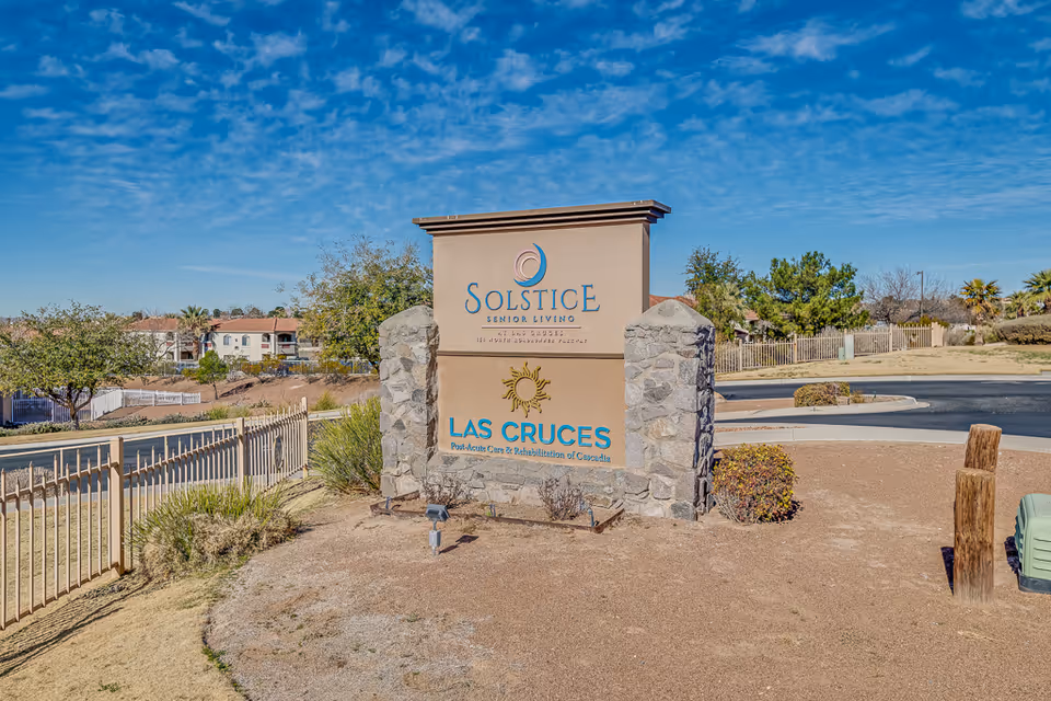 Stone entrance sign for Solstice Senior Living at Las Cruces on a landscaped lot with a road and buildings in the background.