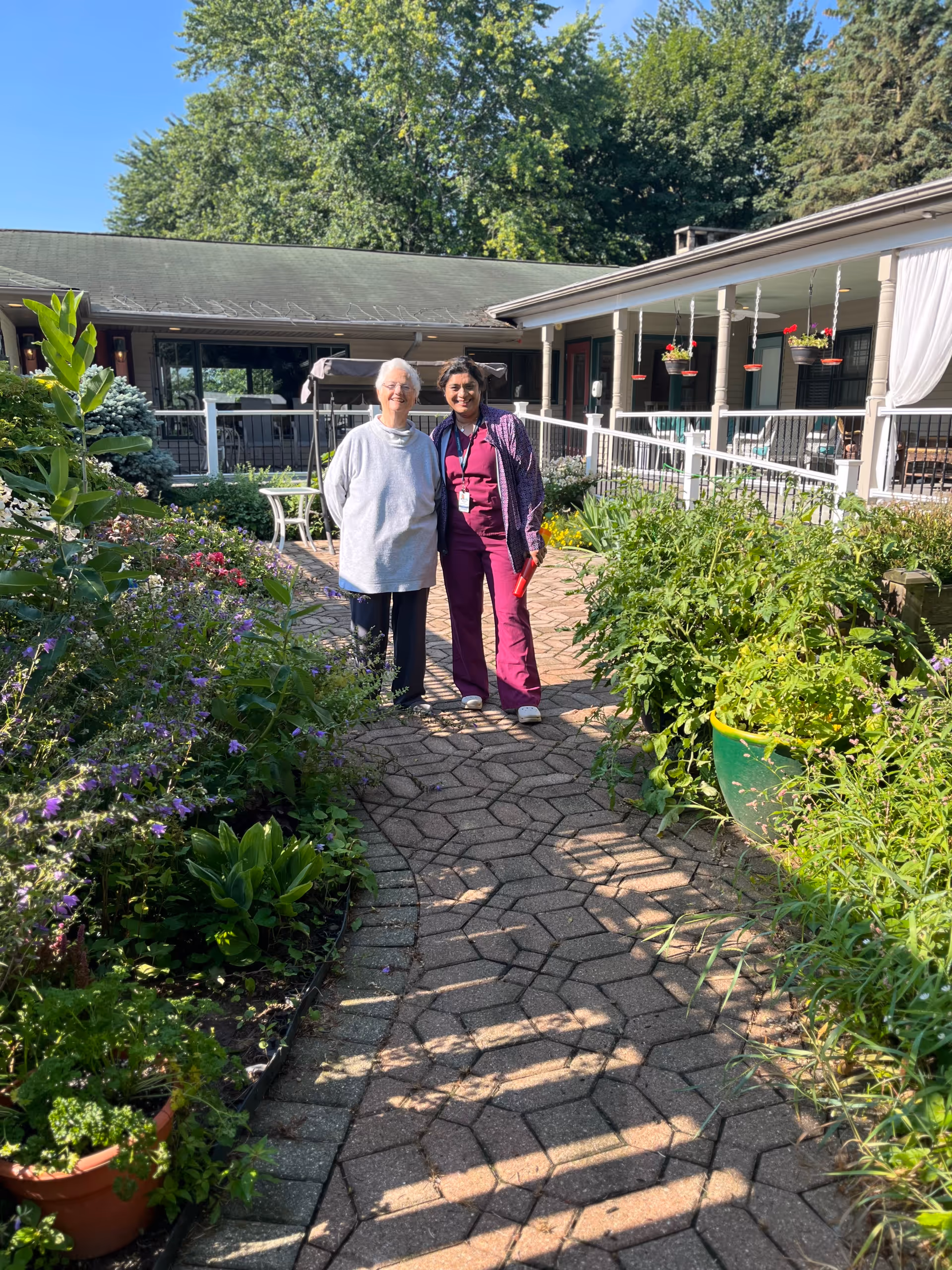 Two women standing on a brick walkway in a lush courtyard garden in front of a single-story senior living building.