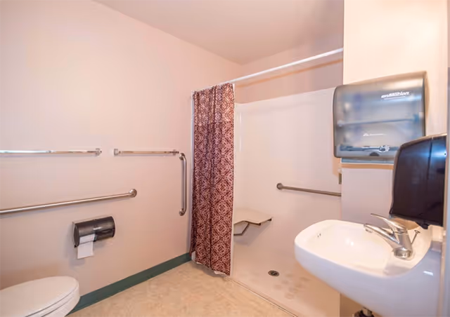 Accessible bathroom with a toilet, a wall-mounted sink, a shower area with a red patterned curtain, and multiple grab bars for support. There is a paper towel dispenser and a soap dispenser mounted on the wall.
