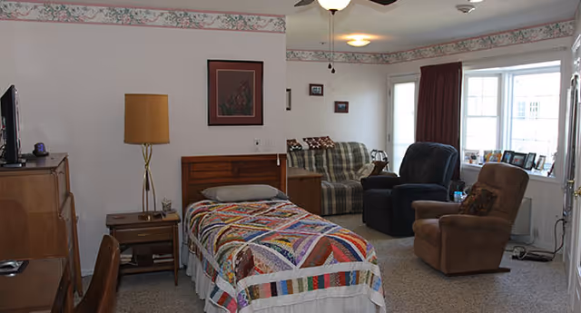 A senior living facility room with a single bed covered by a colorful patchwork quilt, a wooden nightstand with a lamp, a wooden dresser with a TV on top, and a seating area with a plaid sofa and two recliner chairs near a window with burgundy curtains.
